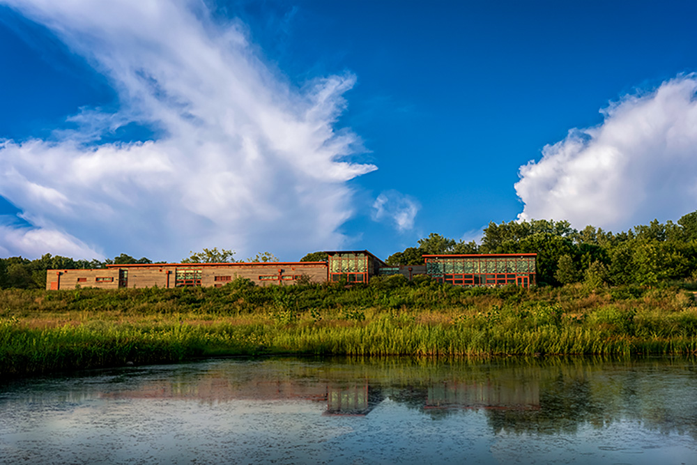 Long, red-brown building reflected in the waters of a marsh against a background of blue sky