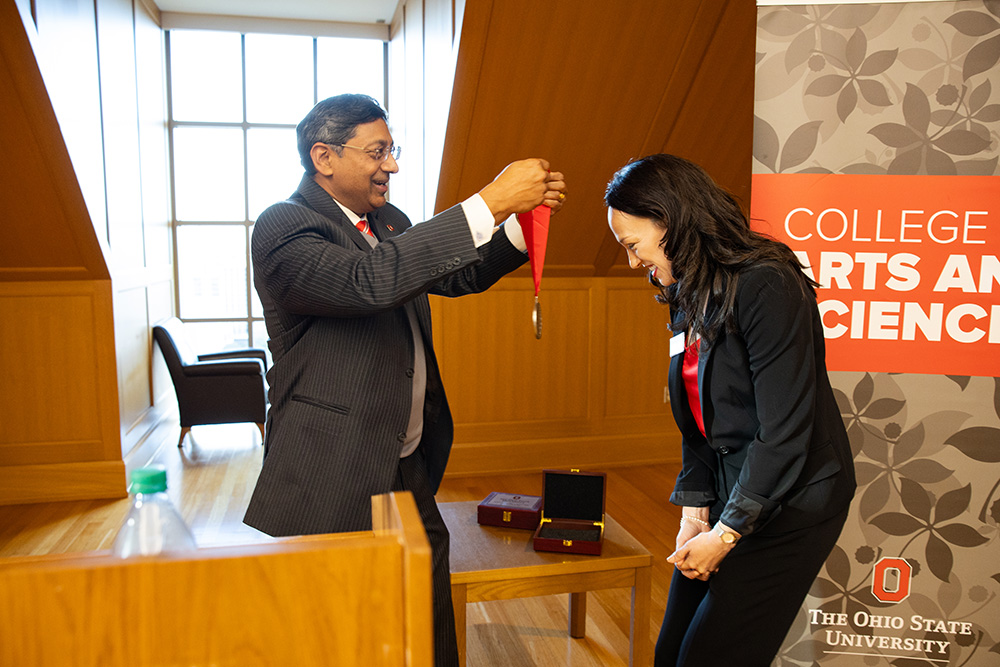 A man in a dark suit prepares to hang a medallion with a red ribbon around the neck of a bowing woman in a dark suit.