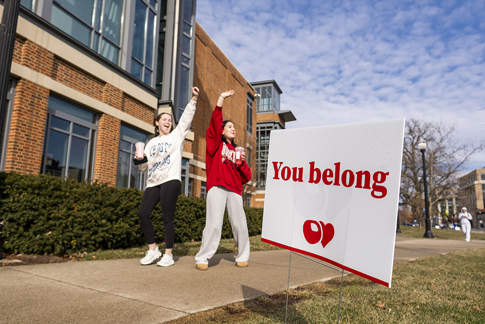 Two students jumping and waving outside of the Union near a white sign that reads "You Belong"