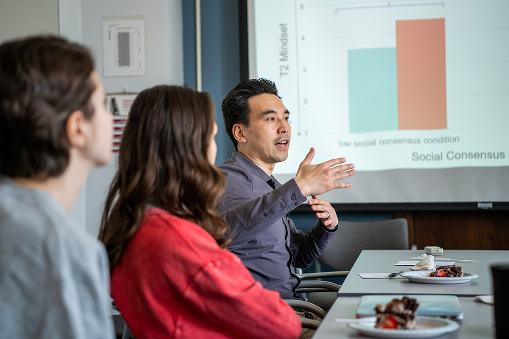 Kentaro Fujita speaks to students seated around a table while a graph is displayed on the screen behind him.
