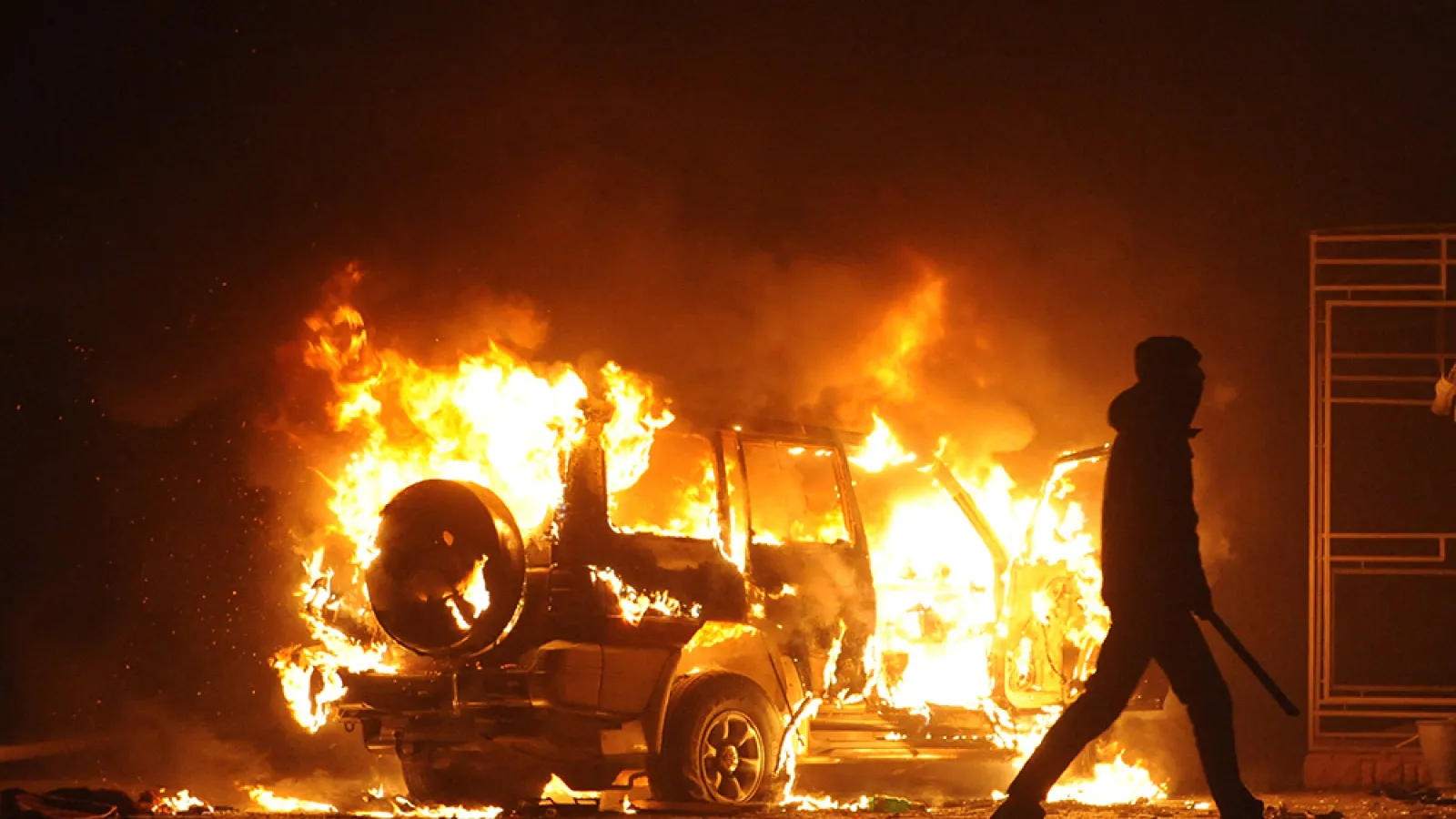 A silhouetted figure carrying a baton walks away from a burning car.