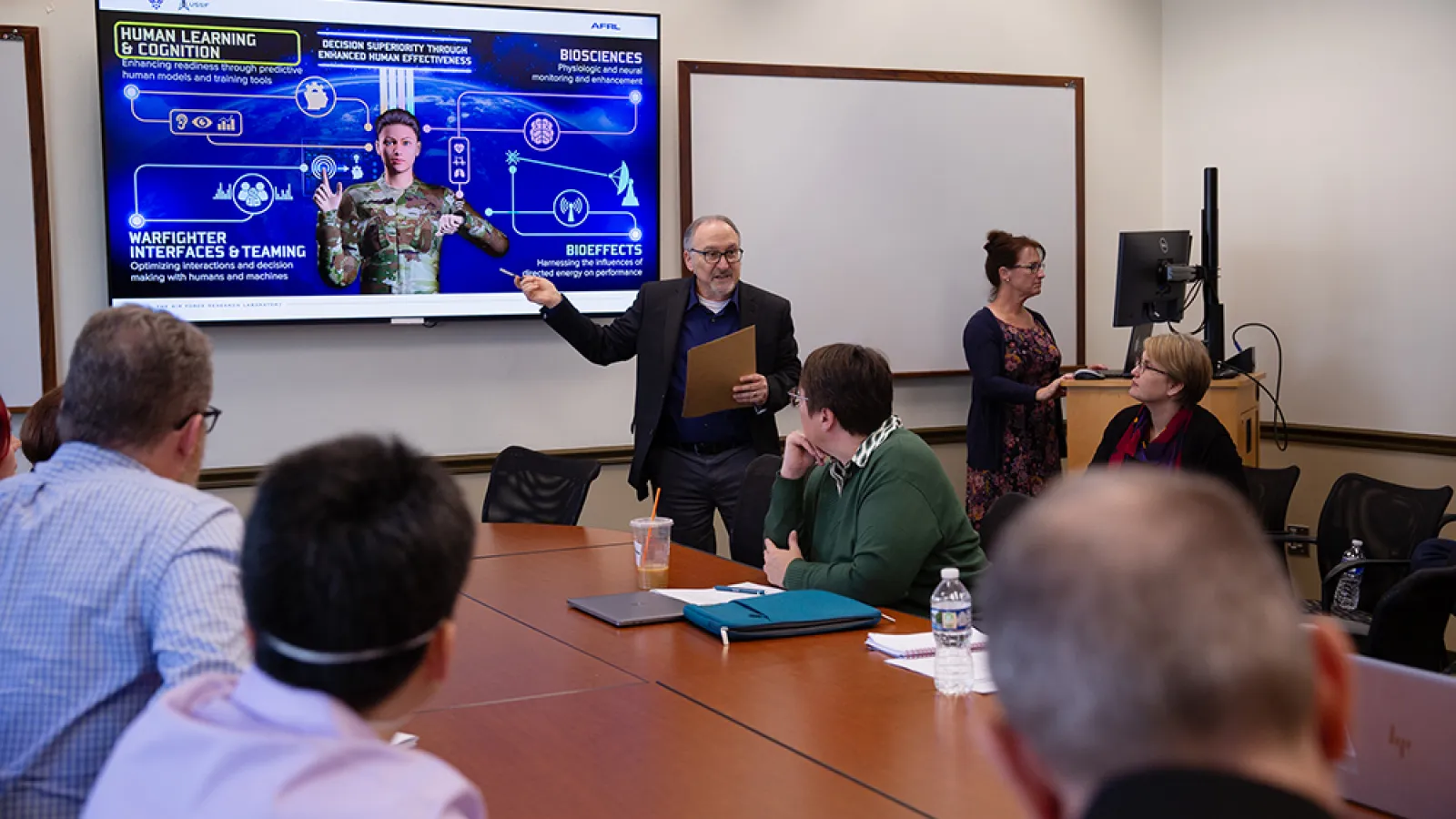 A man points to an image of an armed forces member on a projector screen in a full conference room.