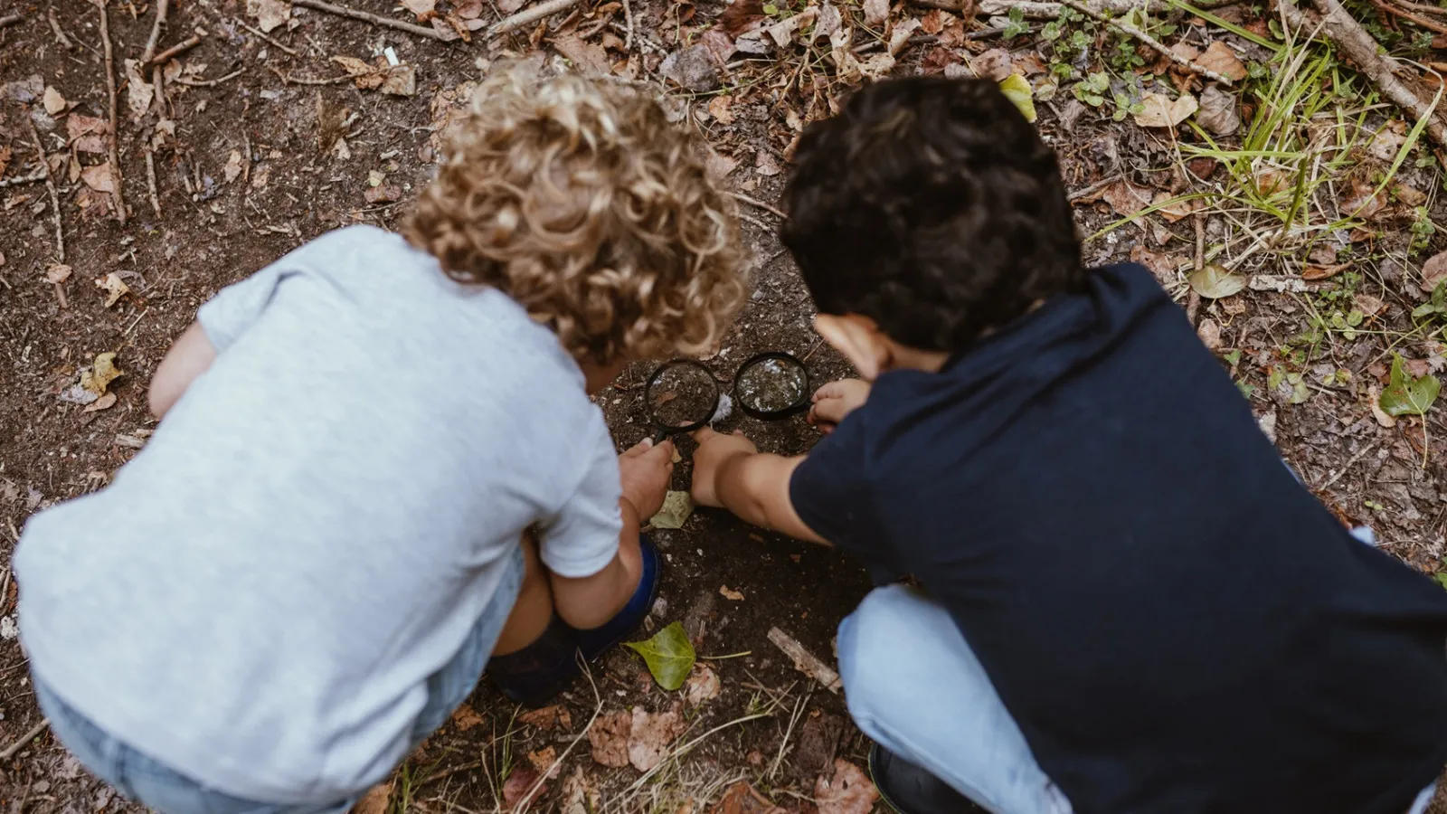 Two children use magnifying glasses to examine dirt on the ground.