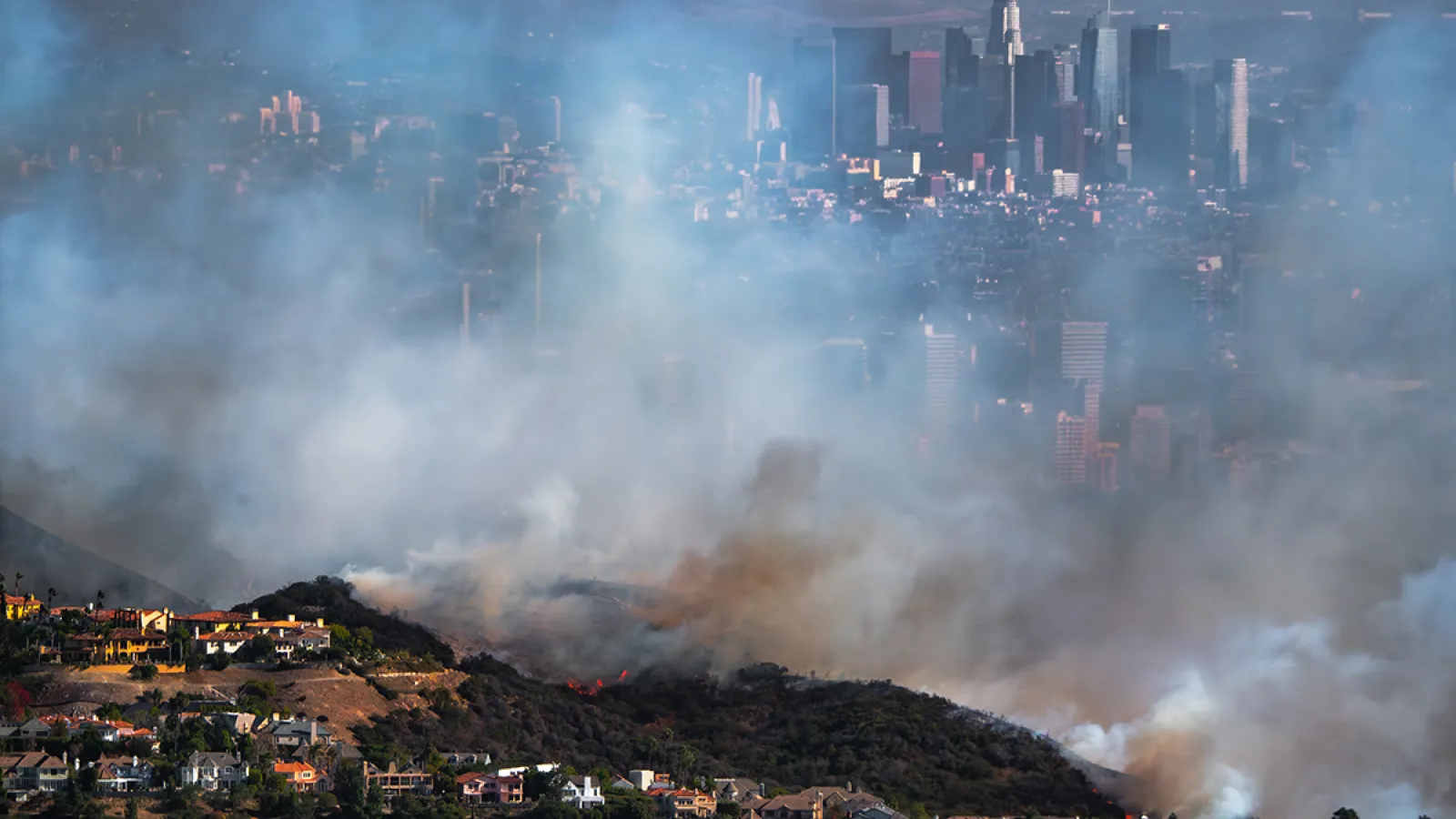 Wildfire smoke looms over a line of hilltop houses with the city of Los Angeles visible in the background