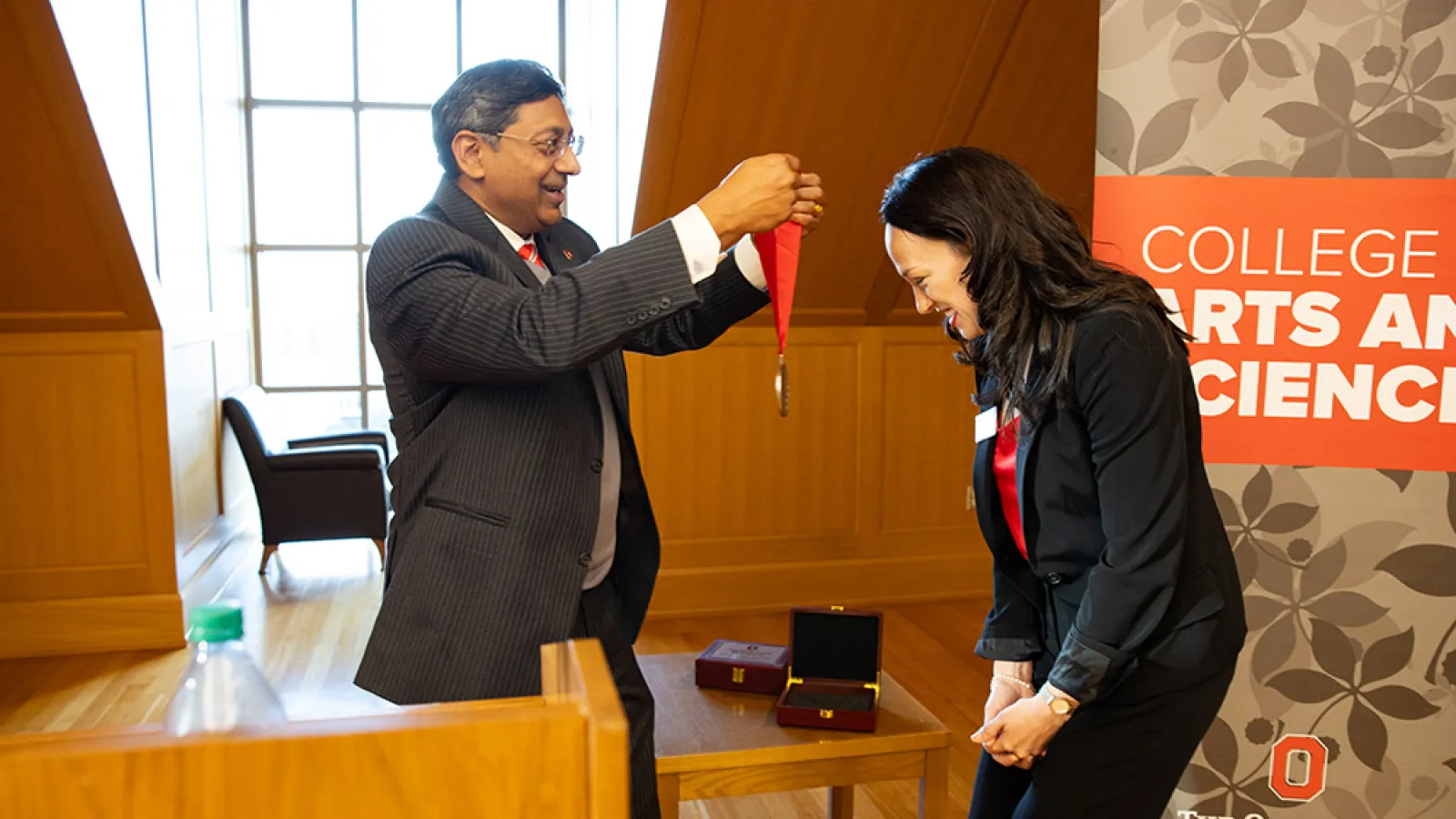 A man in a dark suit prepares to hang a medallion with a red ribbon around the neck of a bowing woman in a dark suit.