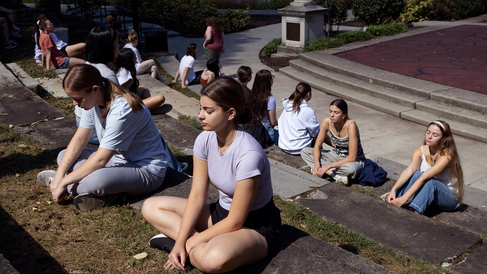 Students sit in rows in an outdoor amphitheater with their eyes closed