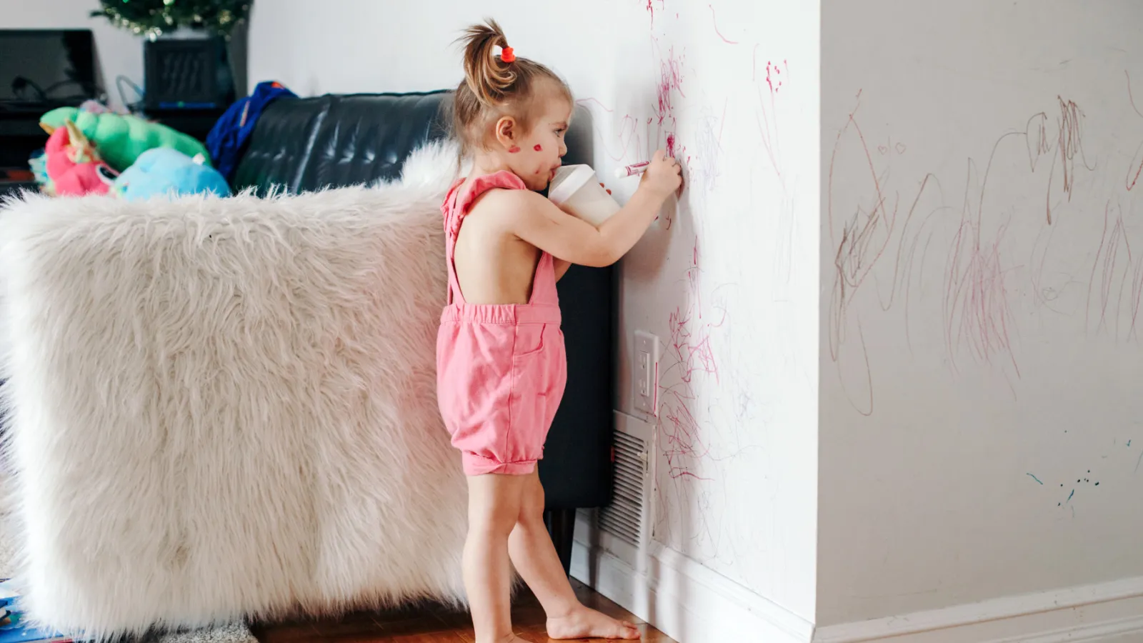 A child scribbles on a while wall using red crayon