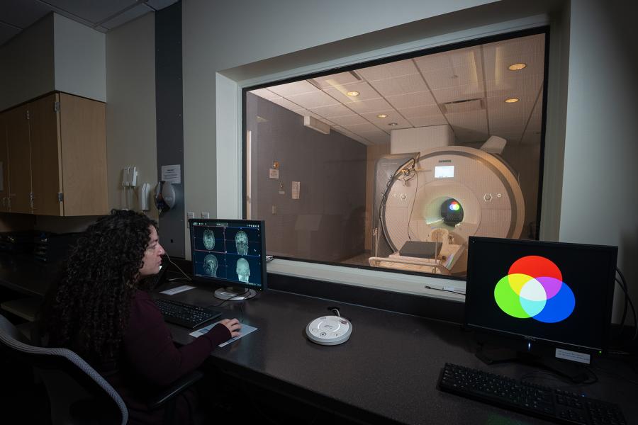 A woman watches a computer monitor with imagery of a human brain as an fMRI machine, visible through the window in front of her, operates.