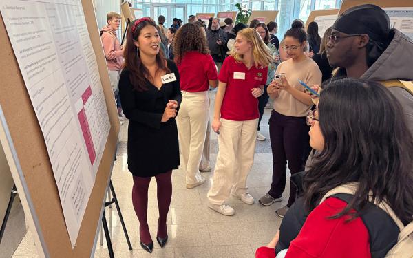 A student researcher speaks to a group of people gathered around her poster in a crowded building lobby.