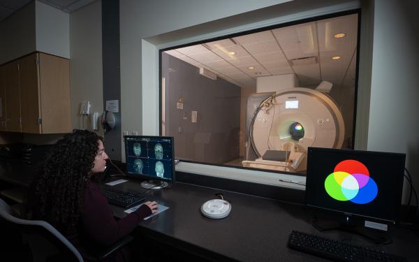 A woman watches a computer monitor with imagery of a human brain as an fMRI machine, visible through the window in front of her, operates.