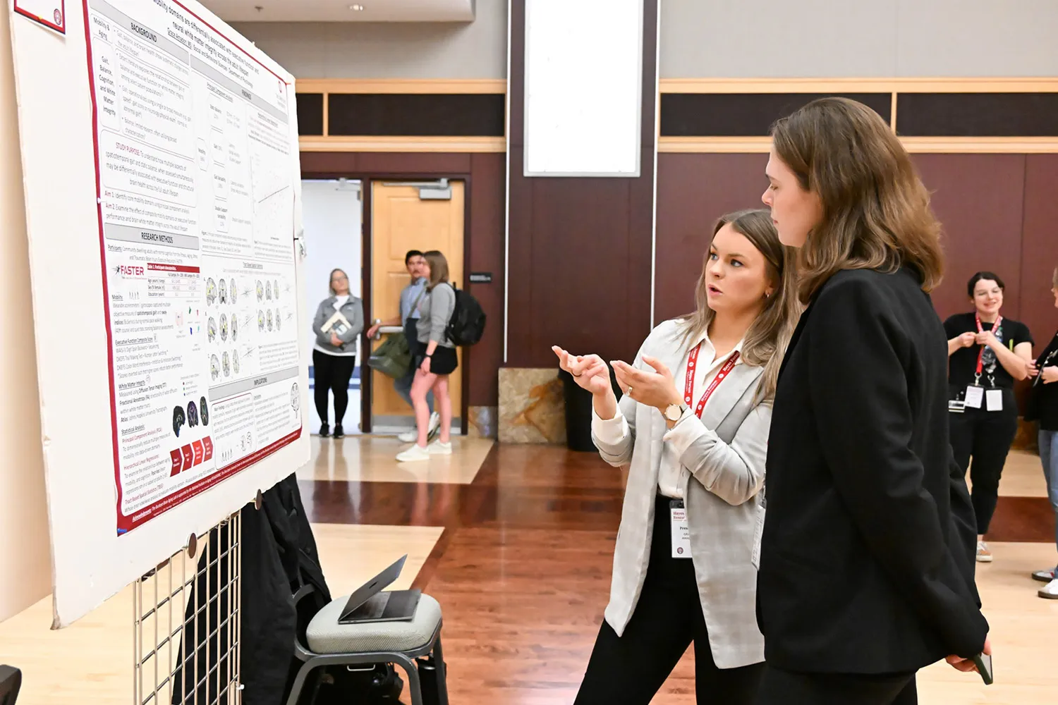 A student presents a poster to a passer-by in an auditorium